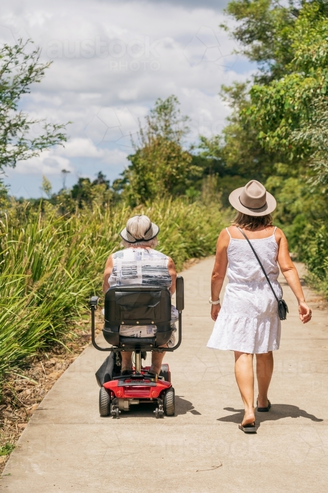 Rear view of an elderly woman on a disability scooter walking with her daughter on a path in a park - Australian Stock Image