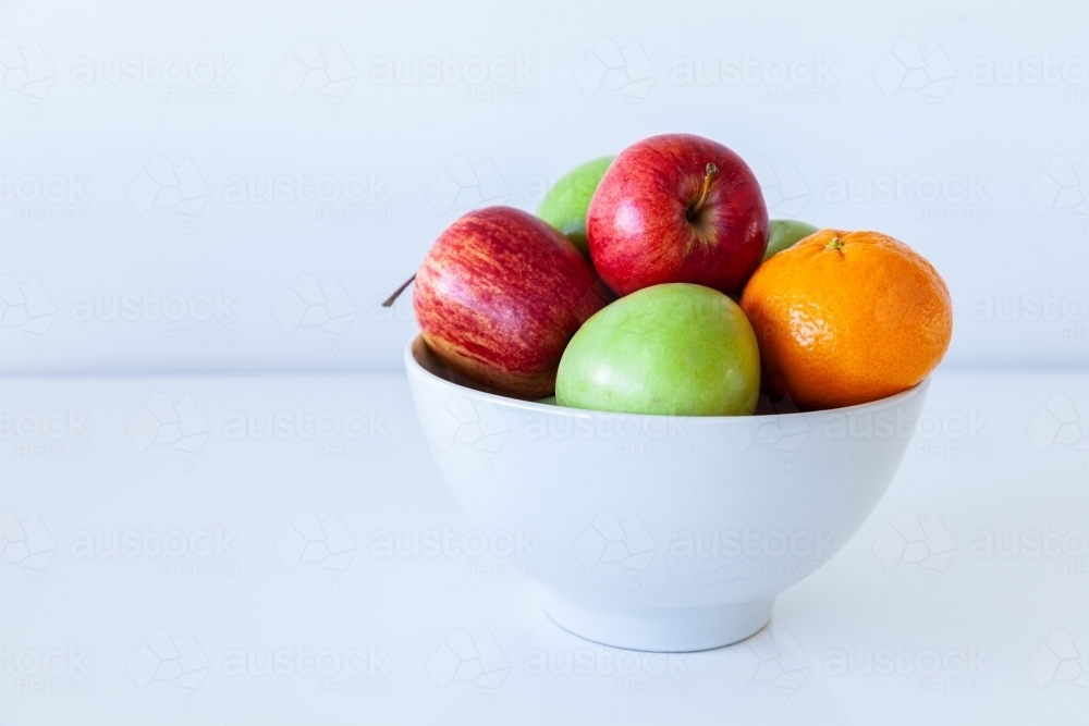 Red and green apples and a mandarin in fruit bowl on white - Australian Stock Image