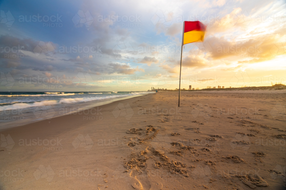 Red and yellow lifeguard flag blowing in wind on sandy beach with tide out and sunset sky - Australian Stock Image
