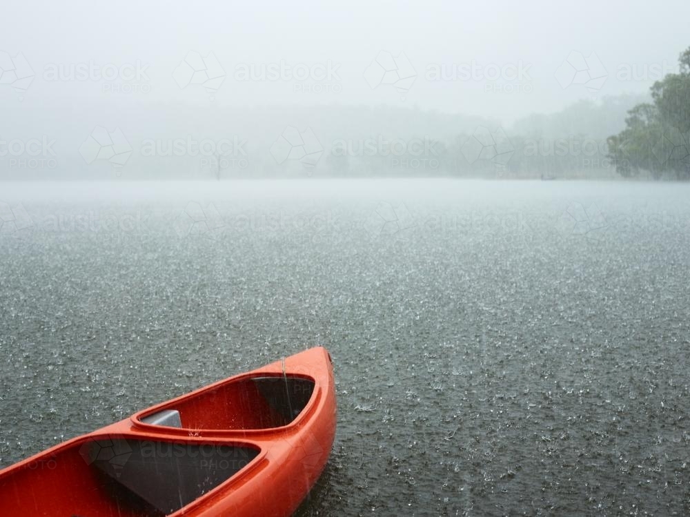 Red canoe sits idle on grey rainswept lake. - Australian Stock Image
