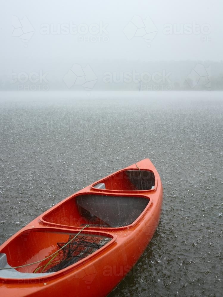 Red canoe sits idle on grey rainswept lake. - Australian Stock Image