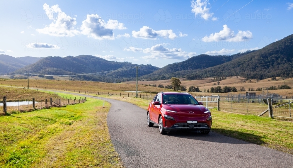 Red electric vehicle car driving down rural country road on sunlit day - Australian Stock Image