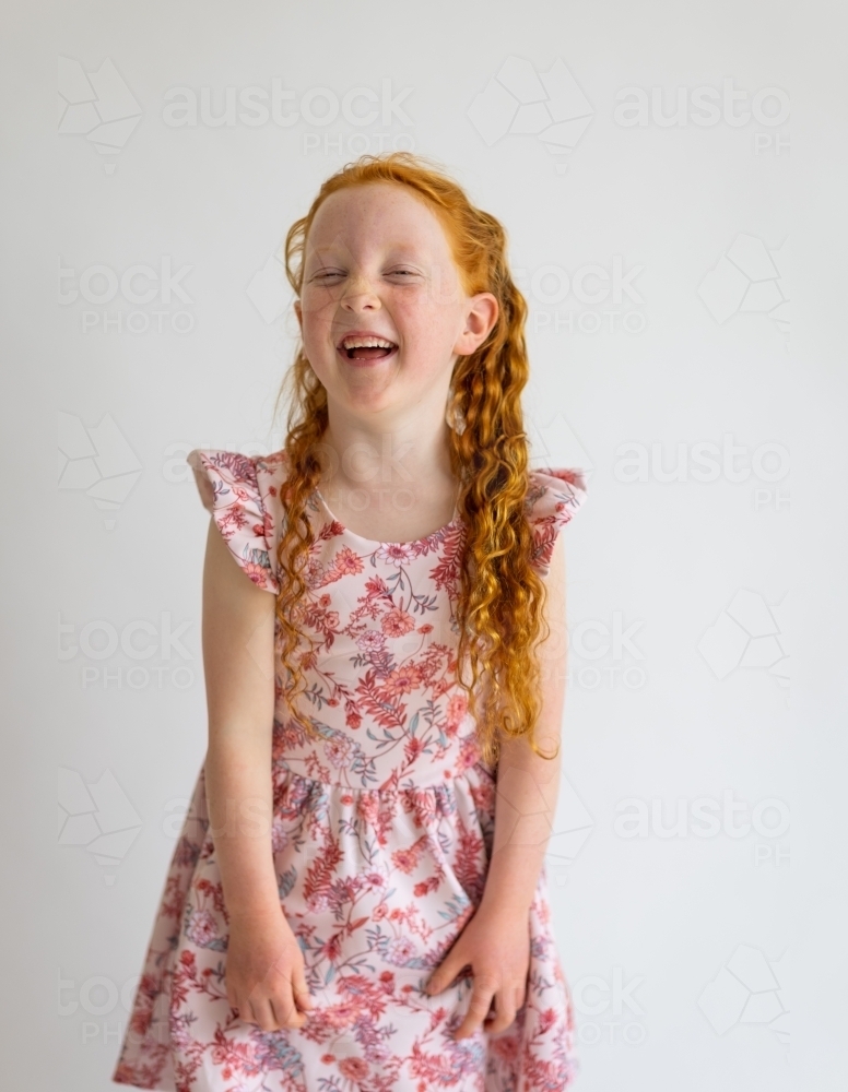 red haired child in floral dress laughing with squinted eyes against plain white background - Australian Stock Image