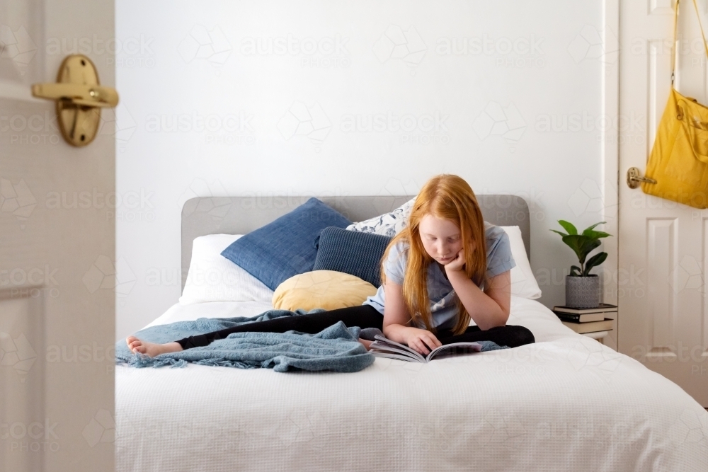 Red haired girl on white bed reading - Australian Stock Image