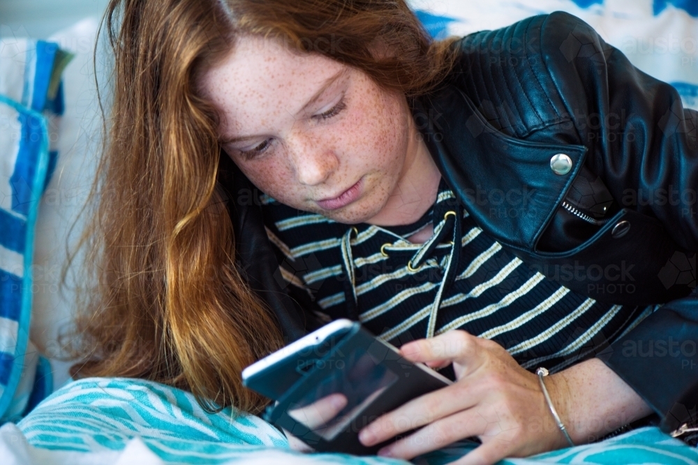 Red haired teenager girl lying on her bed while playing on her phone. - Australian Stock Image