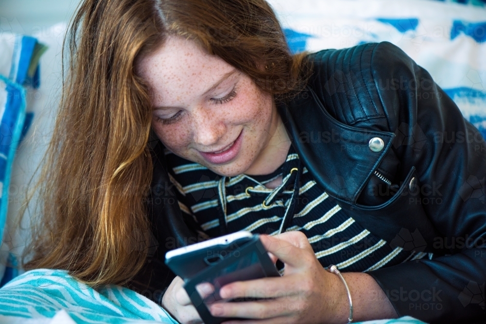 Red haired teenager girl lying on her bed while playing on her phone. - Australian Stock Image
