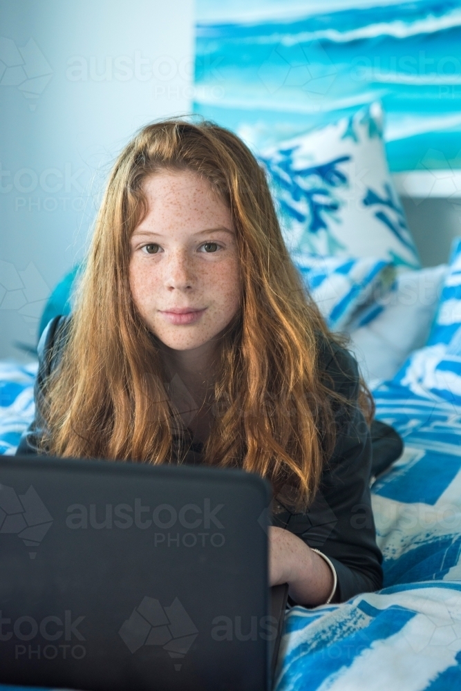 Red haired teenager working on the computer while lying on the bed. - Australian Stock Image