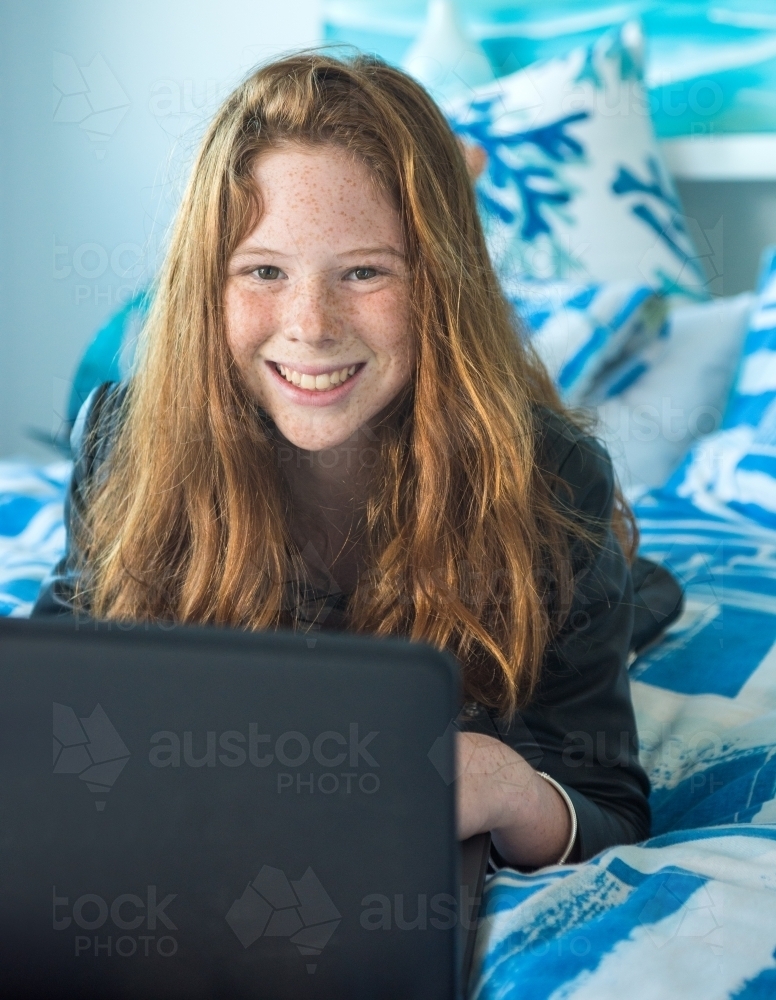 Red haired teenager working on the computer while lying on the bed. - Australian Stock Image