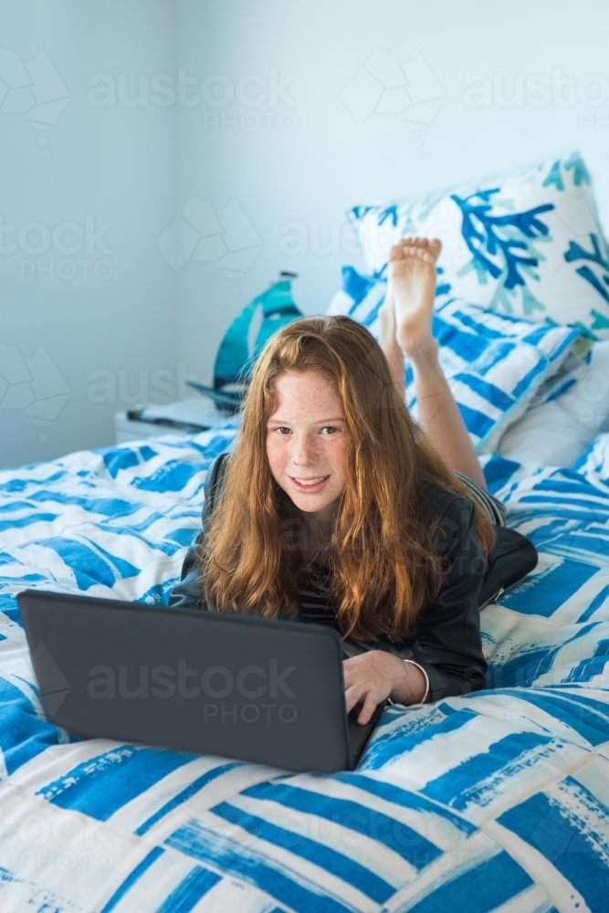 Red haired teenager working on the computer while lying on the bed. - Australian Stock Image