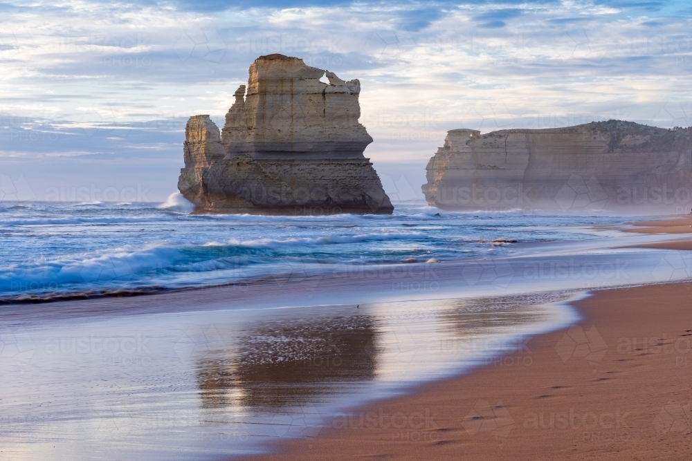 Reflection of a rugged sea stack and cliffs on a wet sandy beach at twilight - Australian Stock Image