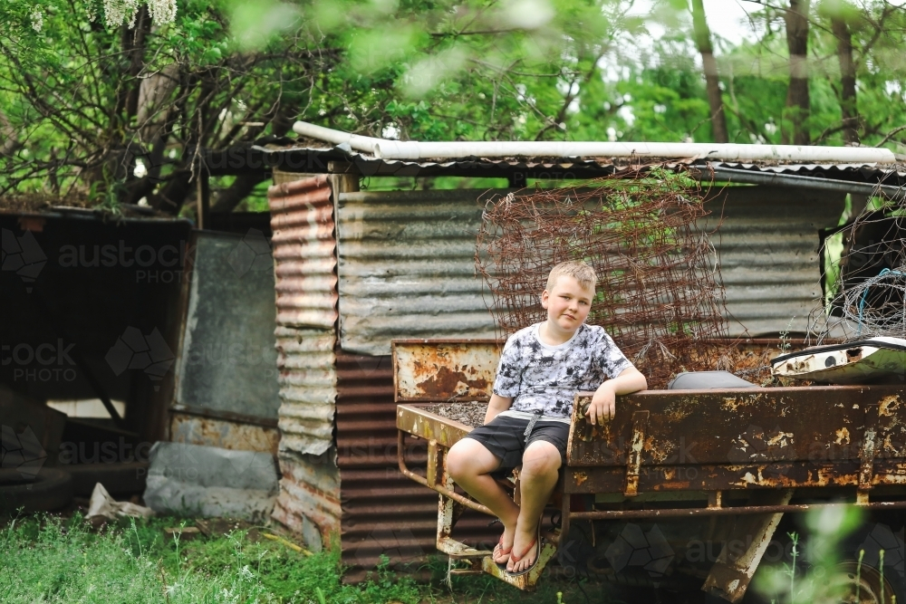 Relaxed boy sitting on ruins of old truck on run down farm - Australian Stock Image