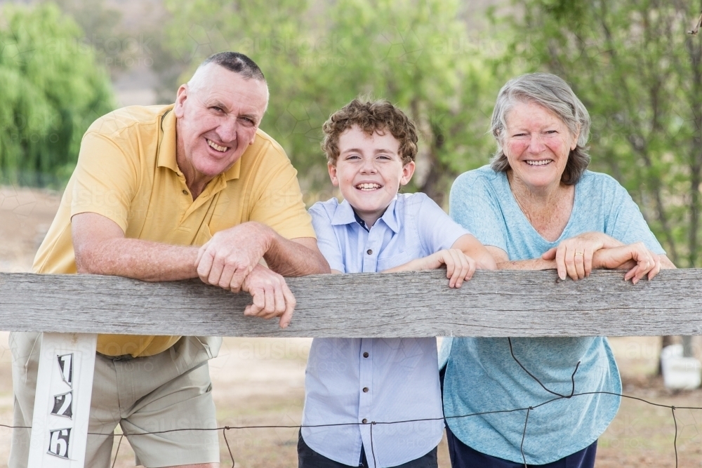 Retired husband and wife grandparents leaning on fence at home happy smiling with grandson - Australian Stock Image