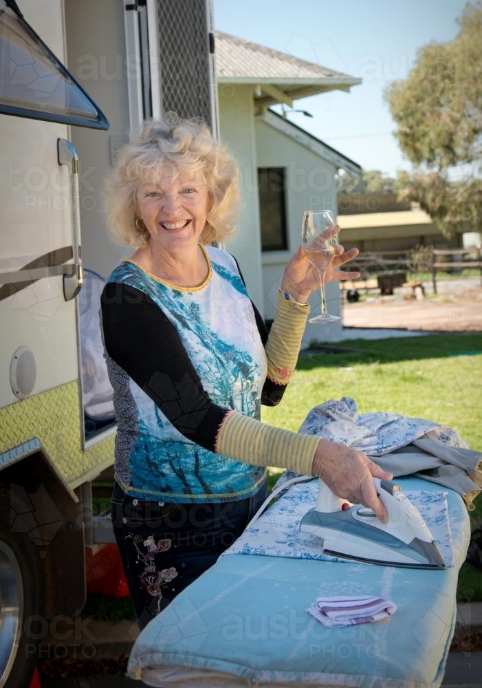 Retired woman ironing outside her caravan with a glass of wine - Australian Stock Image