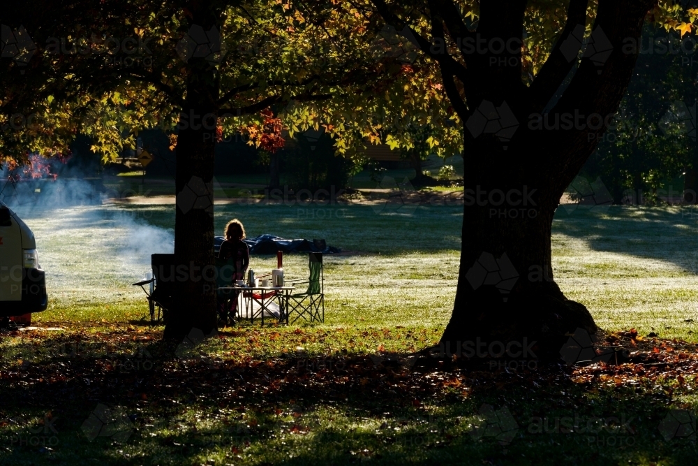 Rim lit silhouette of person sitting at picnic table with smoke under trees with autumn colours - Australian Stock Image