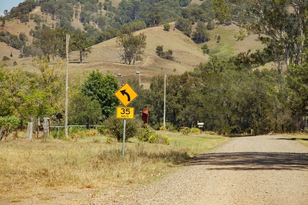 Road sign showing sharp bend ahead recommended speed 35 km per hour on gravel country road - Australian Stock Image
