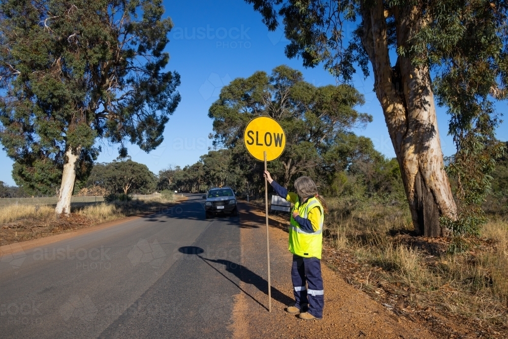 Road traffic controller standing on side of road with slow-stop lollipop sign - Australian Stock Image