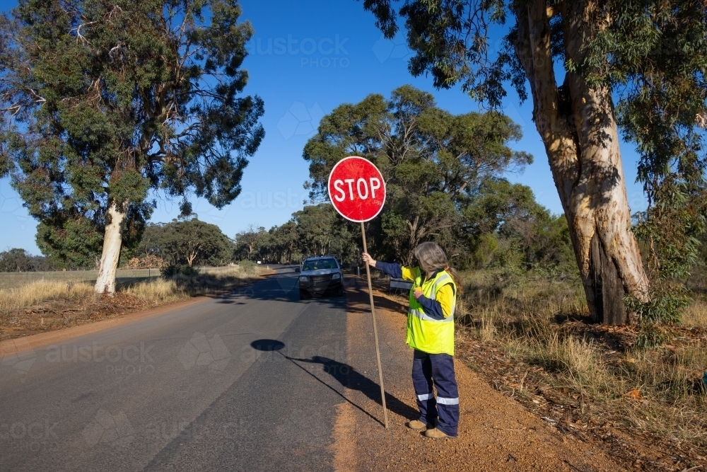 road worker wearing hi-vis workwear holding stop sign on side of road - Australian Stock Image