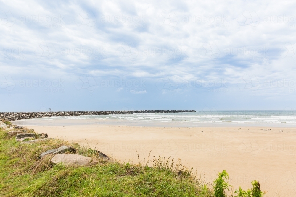 Rock wall curving around beach with sand and water and cloudy skies - Australian Stock Image