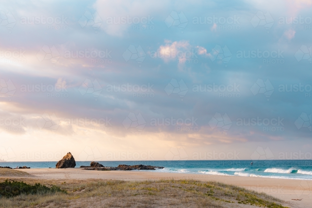 Rocks on Currumbin Beach illuminated at sunset with person windsurf foiling in the distance - Australian Stock Image