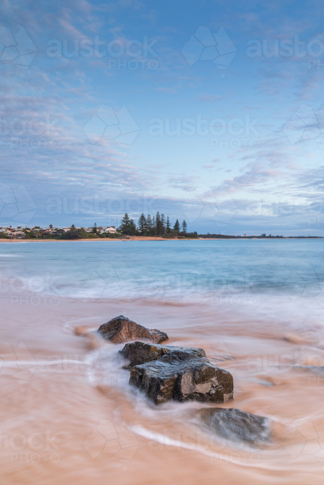 Rocks on golden sand surrounded by ocean with headland in background - Australian Stock Image