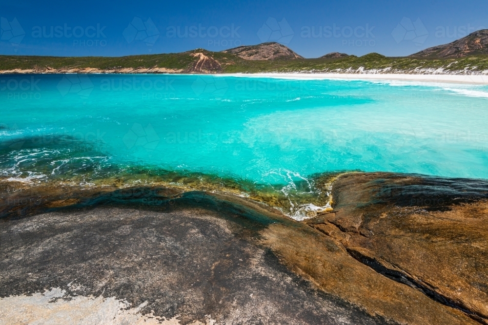 Rocky foreground with clear water and sweeping view of coastline with white sand beach - Australian Stock Image