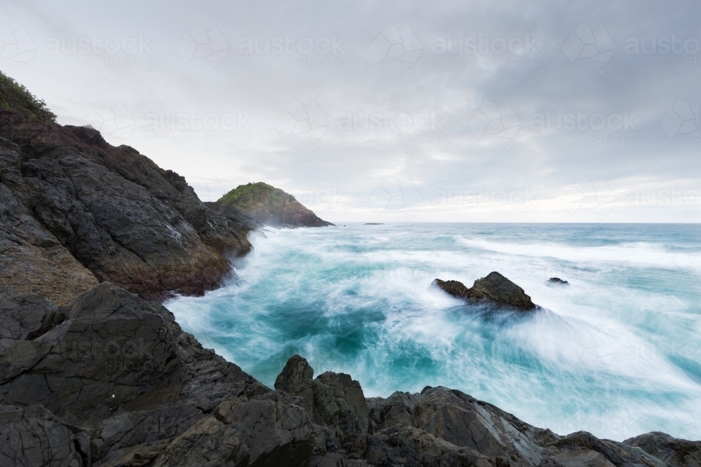 Rough ocean waves crashing against black rocks on an overcast day - Australian Stock Image