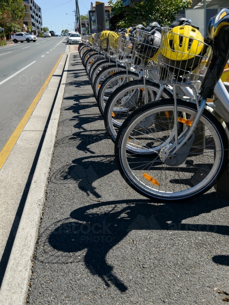 Row of bicycles and helmets with sharp shadows on footpath beside road - Australian Stock Image