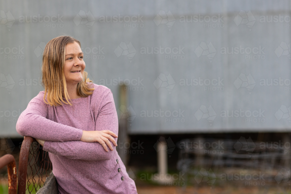 rural woman in her 30s resting her arm on a farm gate near a shed - Australian Stock Image