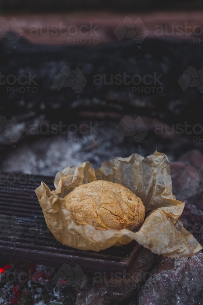 Rustic damper bread in brown baking paper on the red hot coals of a campfire - Australian Stock Image