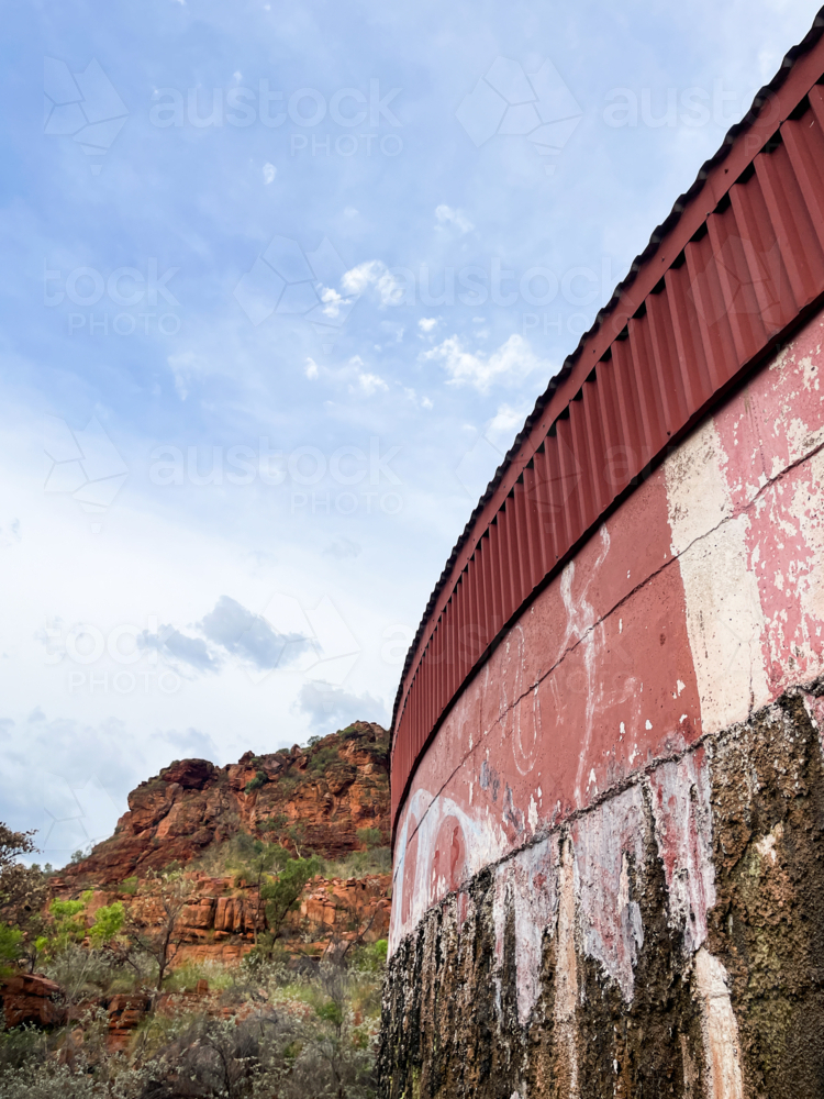 Rustic vandalised water tank with remote rural red rocky ridgeline & blue cloudy sky in background - Australian Stock Image