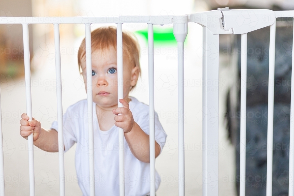 Sad little girl standing at locked baby safety gate and fence - Australian Stock Image