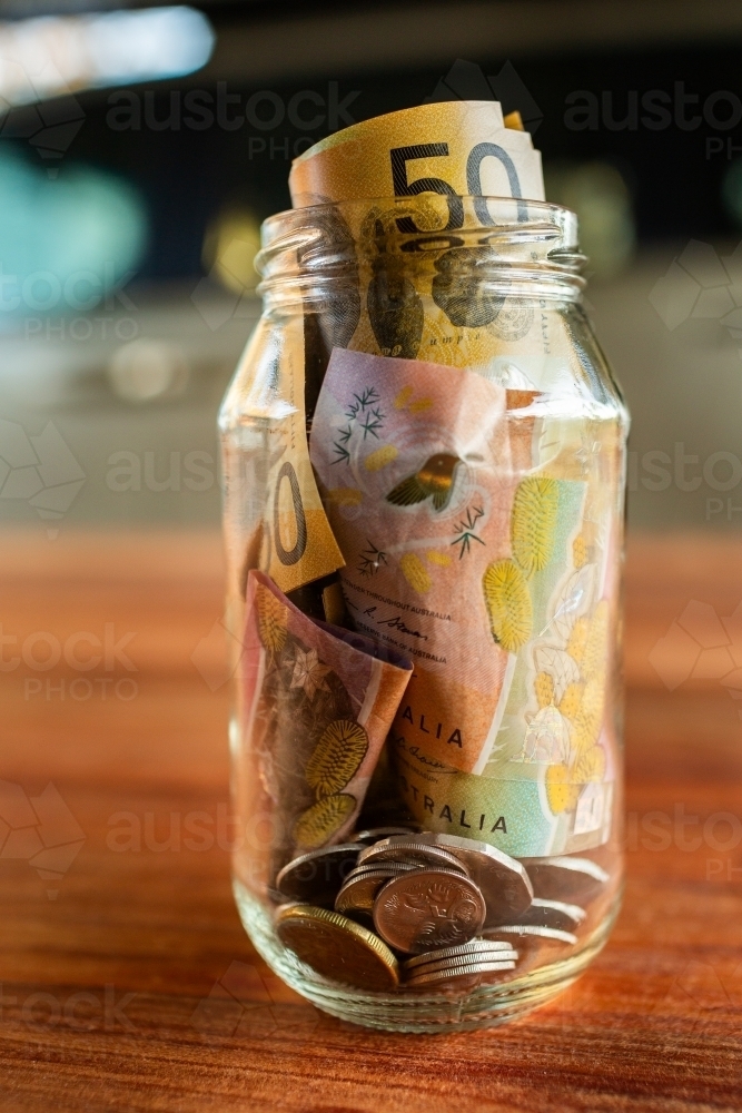 Savings jar of money with coins and fifties in it on table - Australian Stock Image