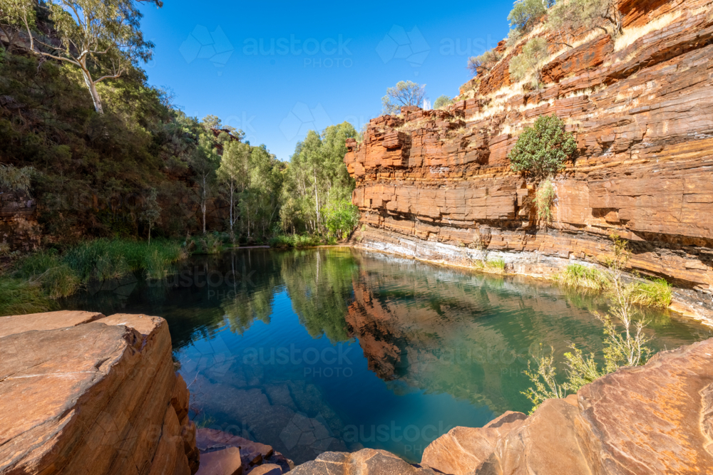 Scenic natural rock pool surrounded by red cliffs and lush vegetation under a clear blue sky. - Australian Stock Image