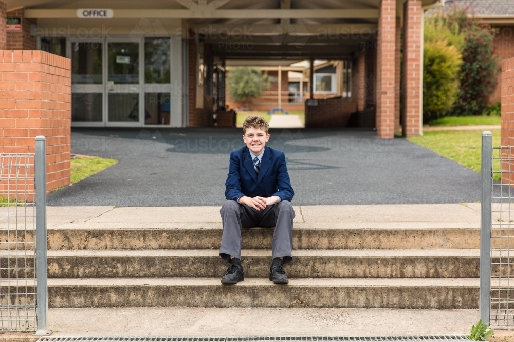 School child in uniform sitting on stairs in front of school office smiling - Australian Stock Image