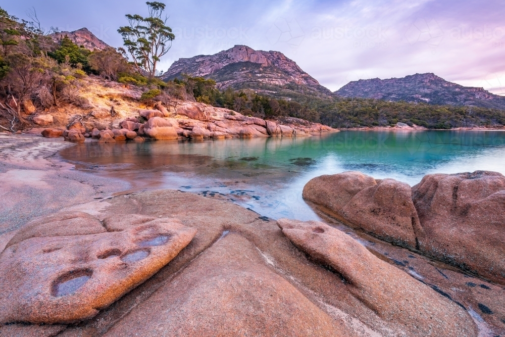 Sculptured granite rocks around a still bay with large mountains behind at twilight - Australian Stock Image