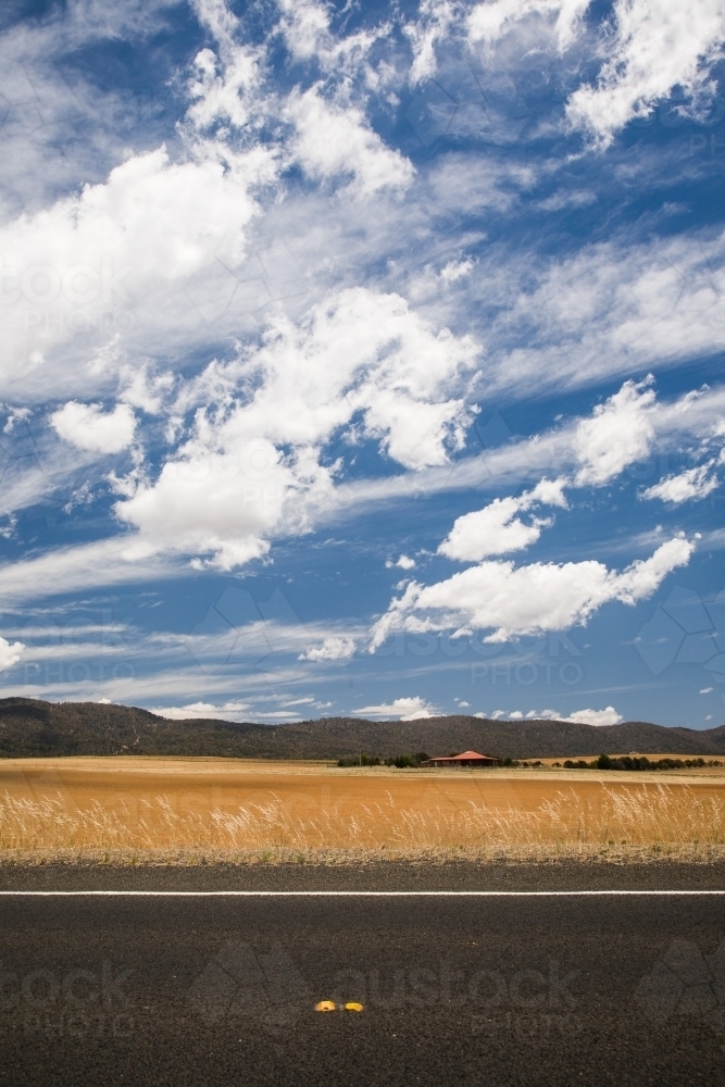 Sealed highway with yellow grass, distant house, blue sky and clouds - Australian Stock Image