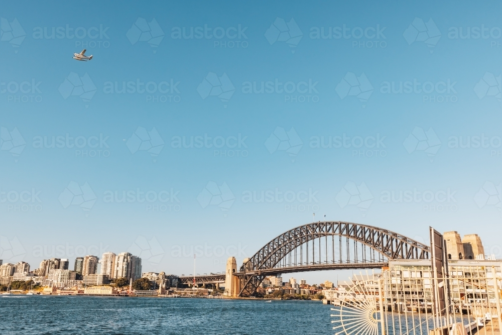 Seaplane flying over the Sydney Harbour Bridge on bright day with blue sky - Australian Stock Image
