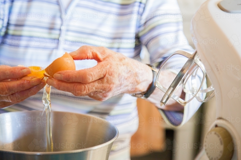 Senior citizen cooking a cake in the kitchen, cracking eggs into a bowl - Australian Stock Image