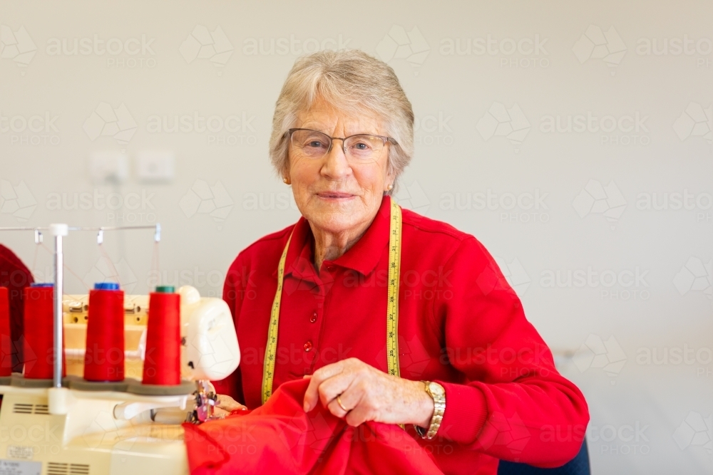 senior lady holding a red fabric on an overlocker machine - Australian Stock Image