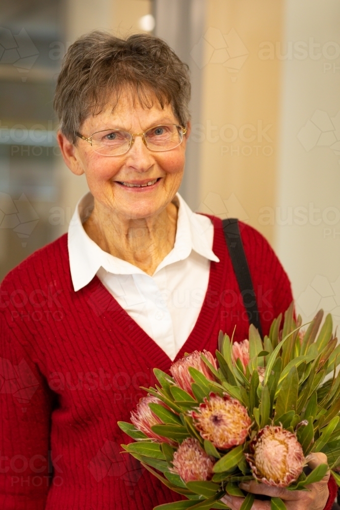 senior lady in dark red cardigan holding bunch of protea flowers - Australian Stock Image