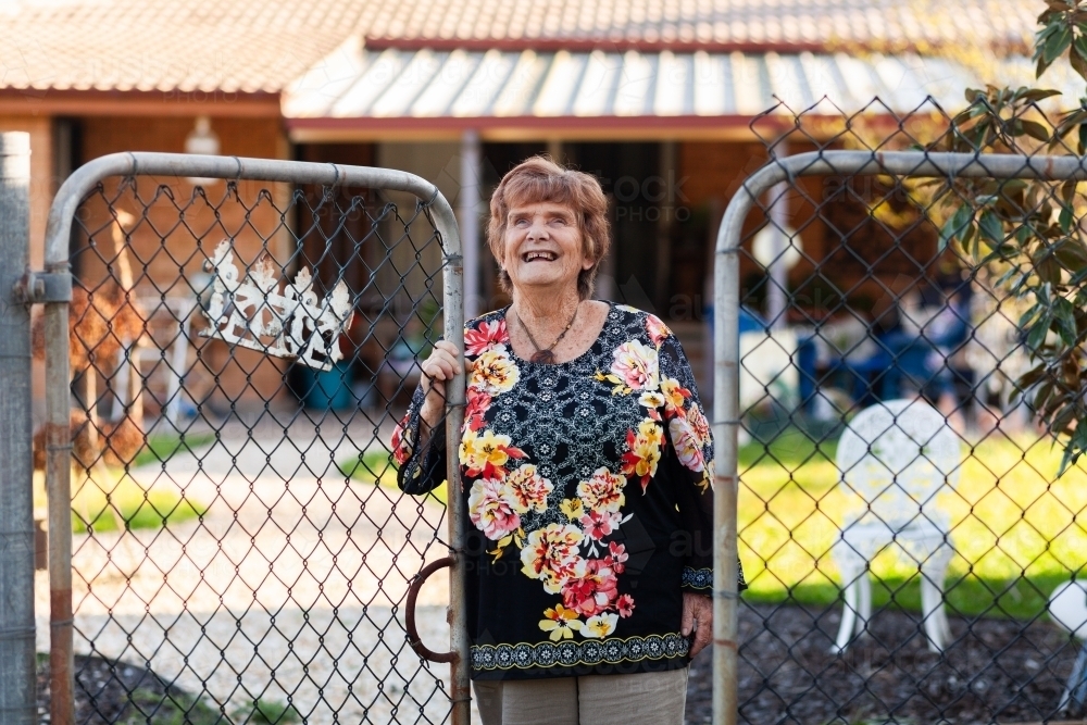 Senior lady looking up smiling while exiting backyard through gate - Australian Stock Image