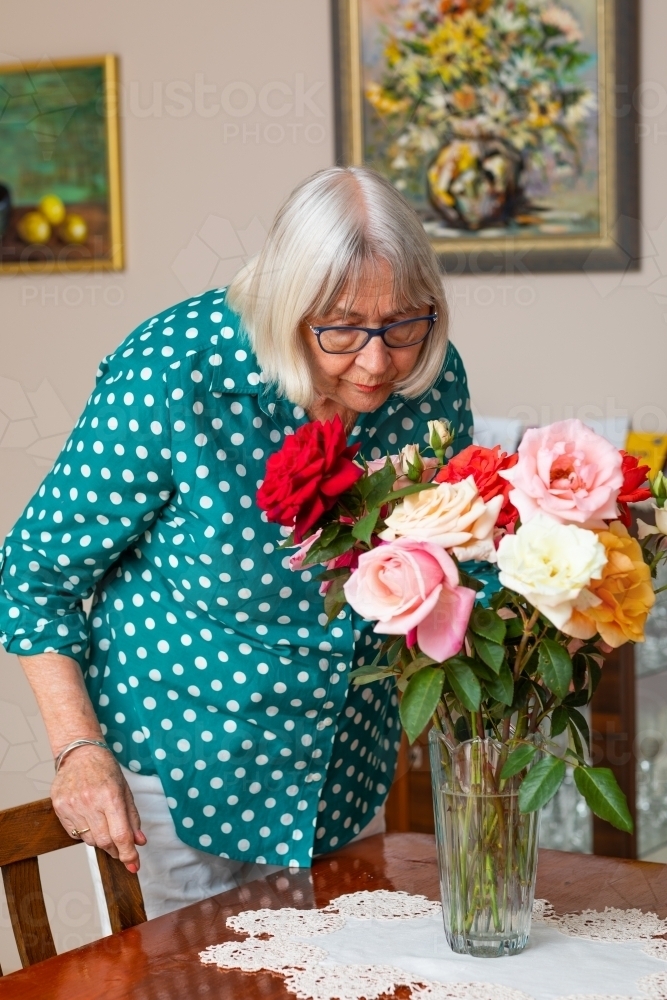senior lady sniffing roses in vase - Australian Stock Image