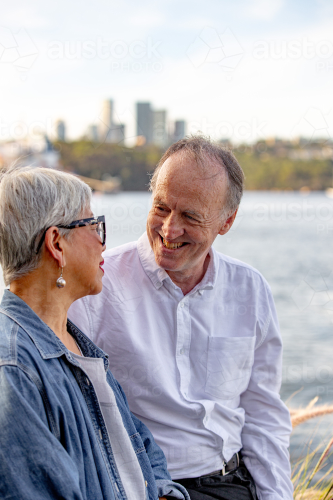 Senior man looking at Asian woman while sitting outdoors near the water : Austockphoto Senior man looking at Asian woman while sitting outdoors near the water - Australian Stock Image
