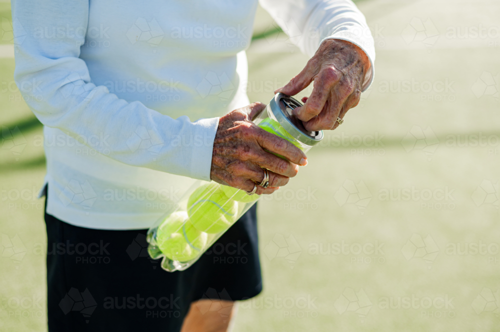 Senior woman prepares for tennis by opening a canister of bright yellow balls outdoors - Australian Stock Image