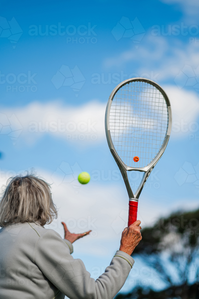 Senior woman serves tennis ball, enjoying a sunny day outdoors - Australian Stock Image