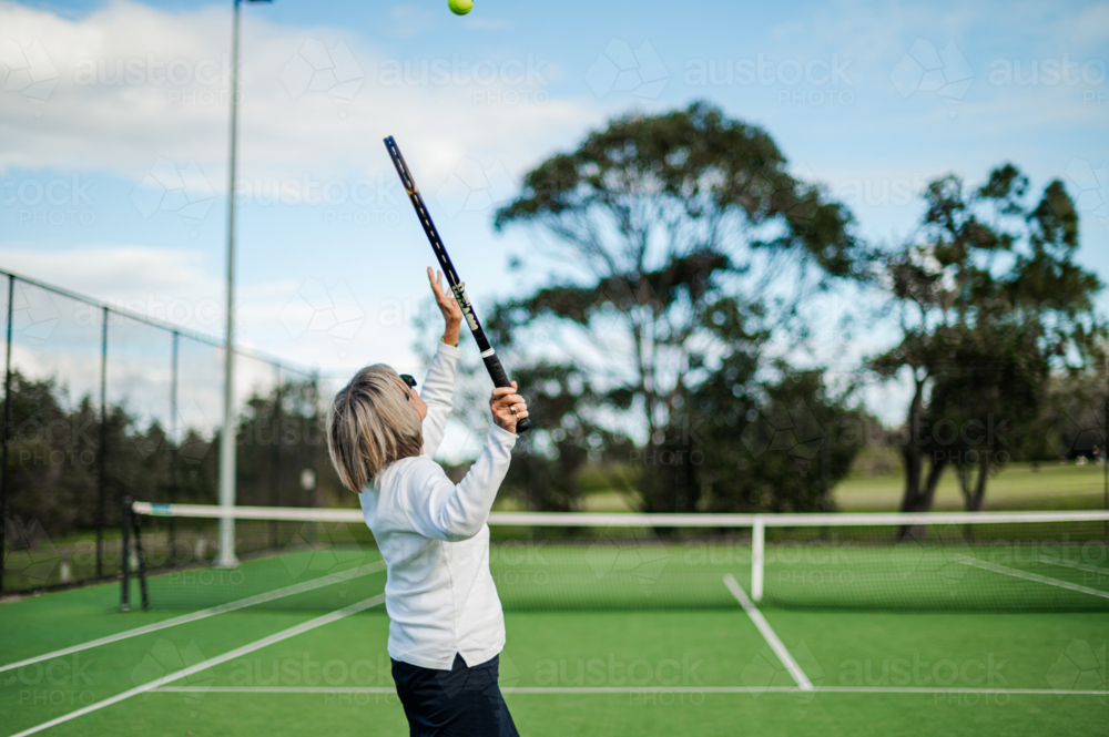 Senior woman serves tennis ball on green court with clear skies - Australian Stock Image