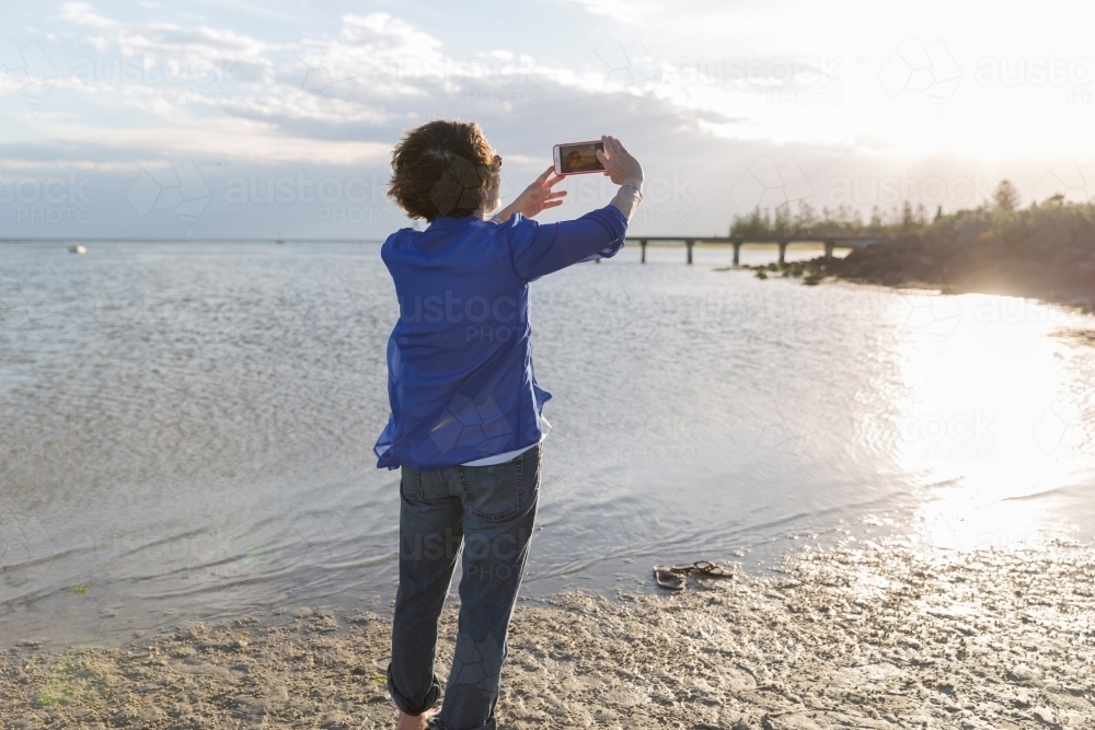 Senior woman taking phone photo on shoreline - Australian Stock Image
