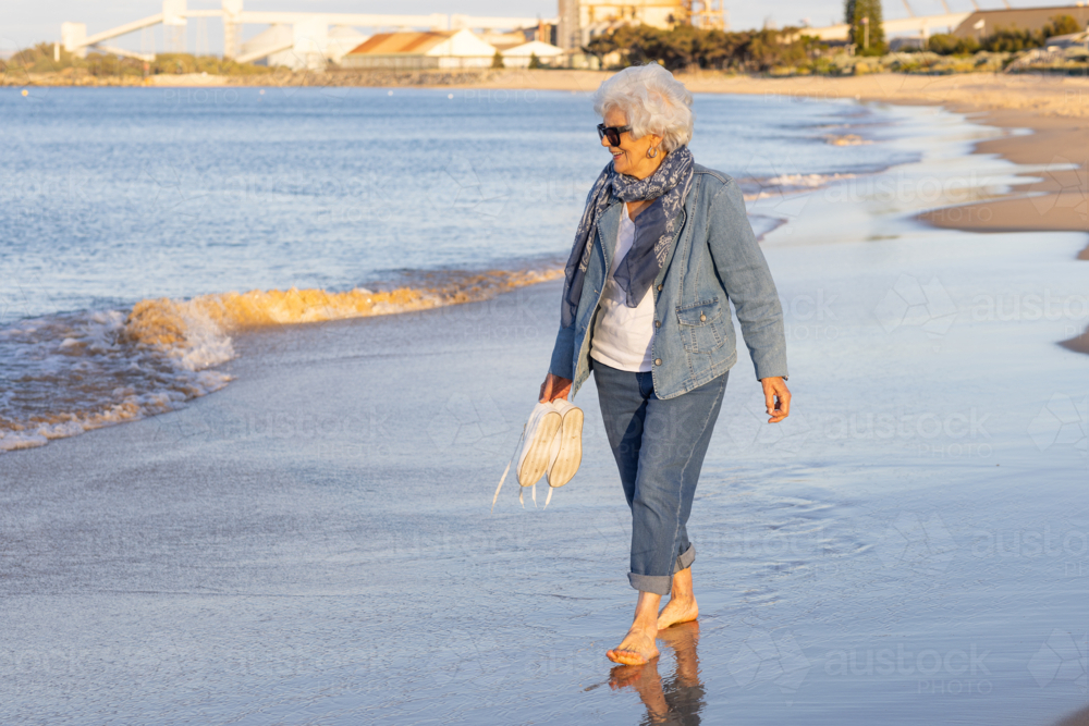 Senior woman walking along the beach barefoot - Australian Stock Image