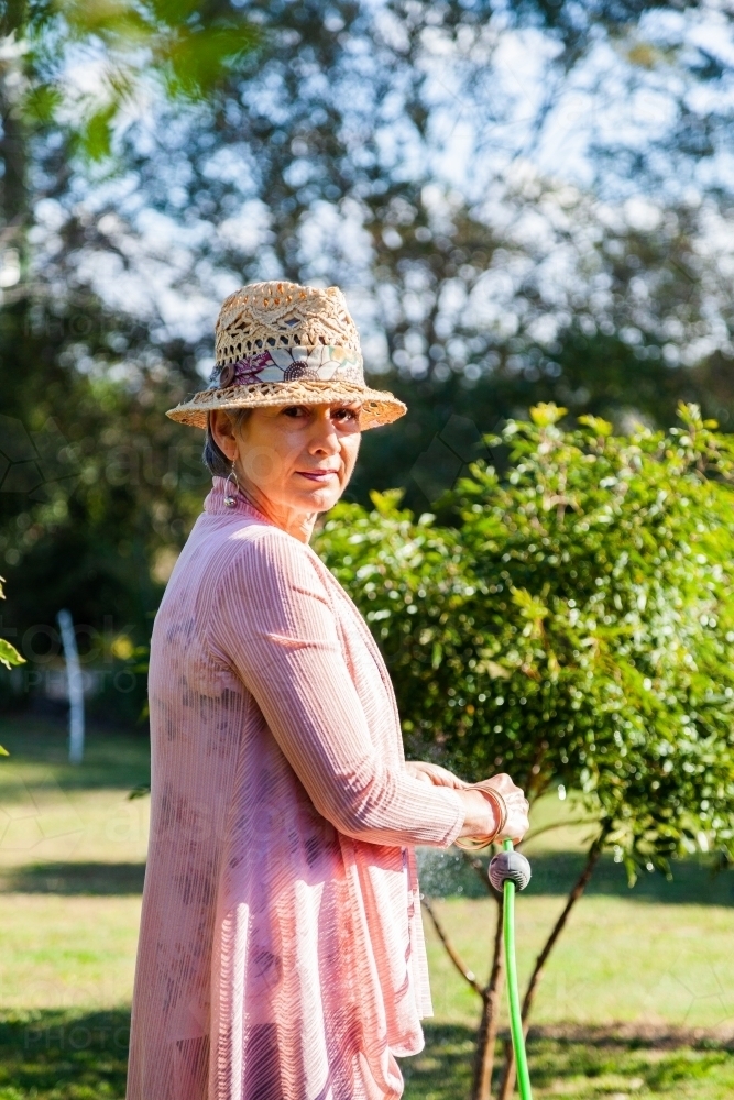 Senior woman watering plants in the garden with green hose - Australian Stock Image