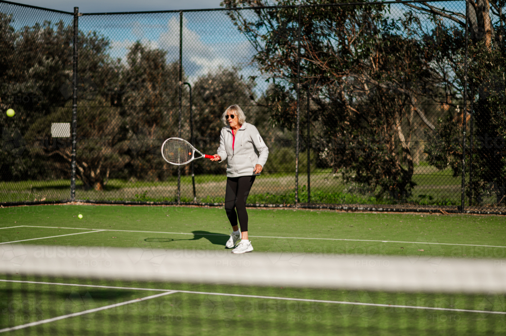 Senior woman wearing sporty attire practices tennis outdoors on court - Australian Stock Image